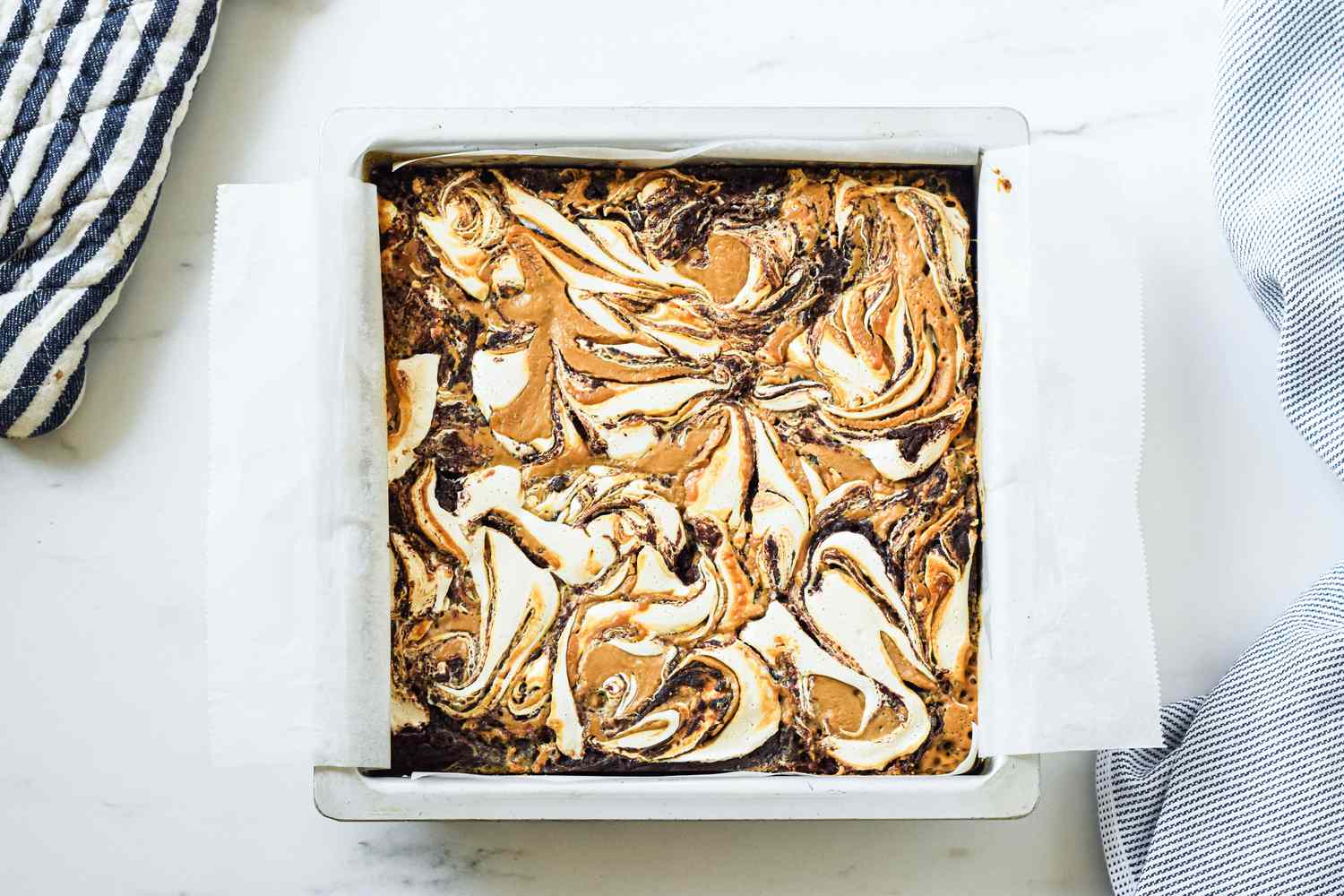Baked fluffernutter brownie in the baking pan and next to it, the blue and white striped kitchen towel and a blue and white striped pot holder