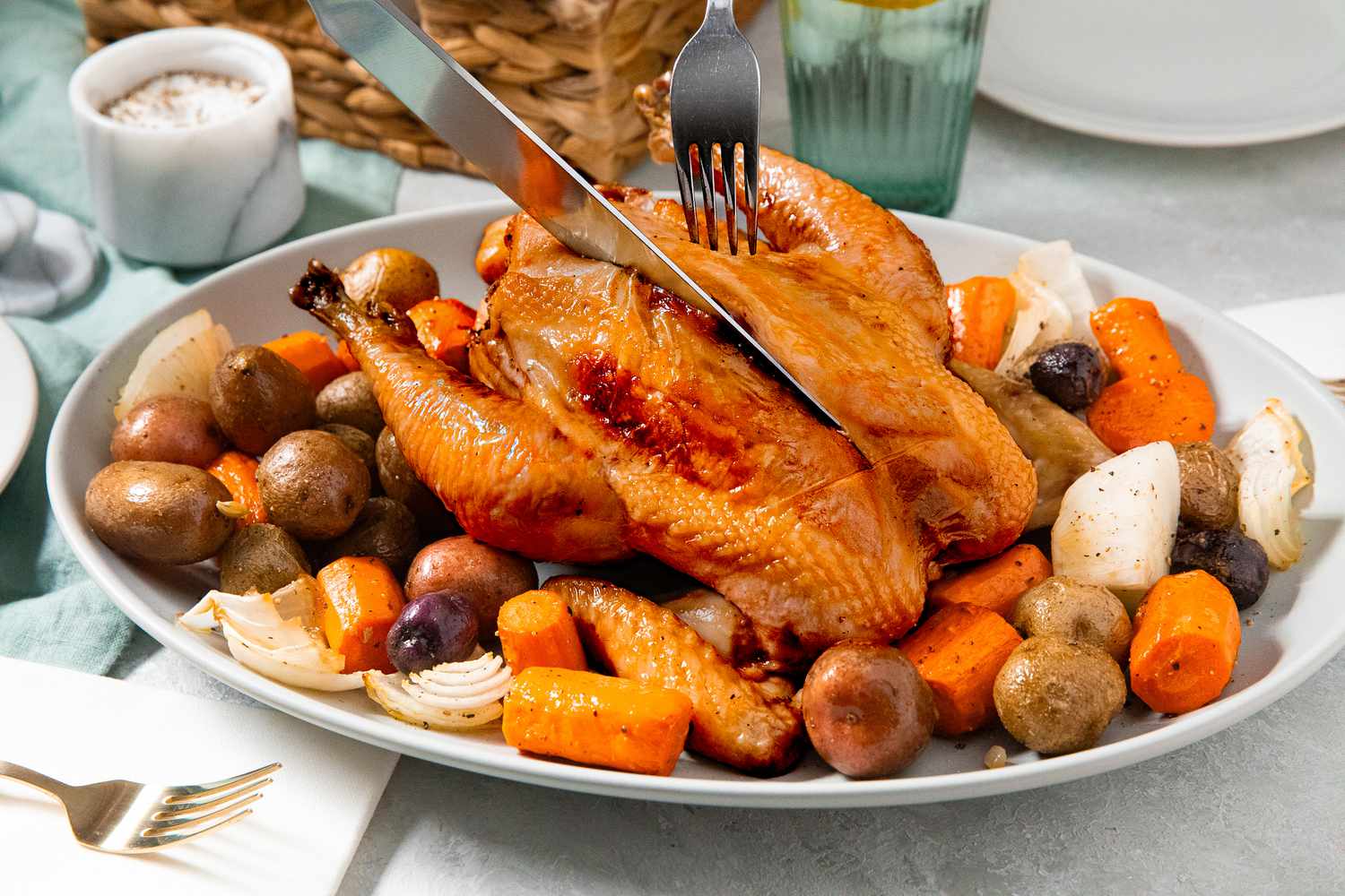 Carving knife cutting into simple roast chicken that's served on a bed of roasted vegetables at a table setting with plates, a bread basket, a glass, and utensils 
